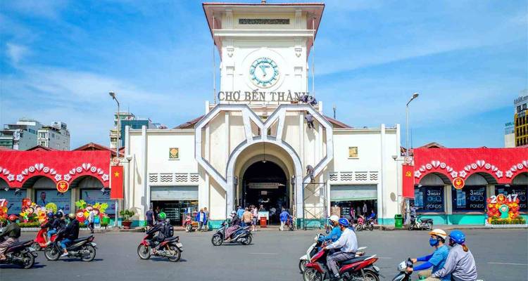 Voorkant van Ben Thanh Market met druk bromfietsverkeer dat er voor langsrijdt onder een heldere hemel.