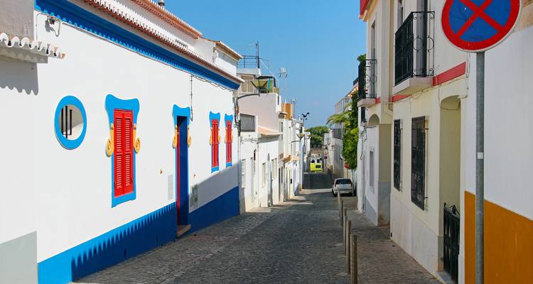 Een smal keienstraatje met witgekalkte huisjes met felblauwe en rode luiken in de Algarve-zon.