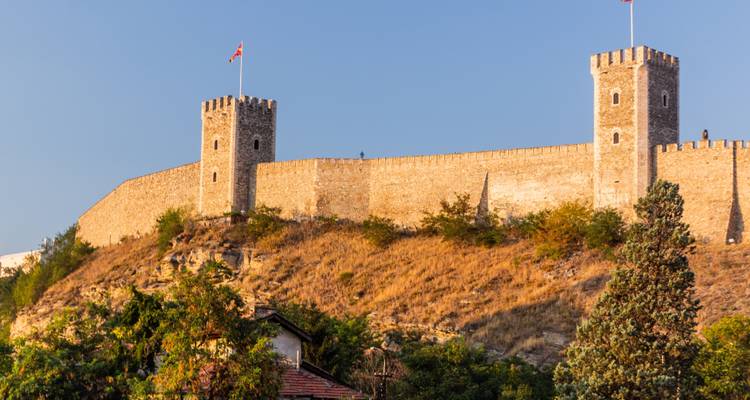 Steinerne Festung mit Zwillingstürmen und einer mazedonischen Flagge, die einen sonnenbeschienenen Hügel über verstreuten Bäumen krönt
