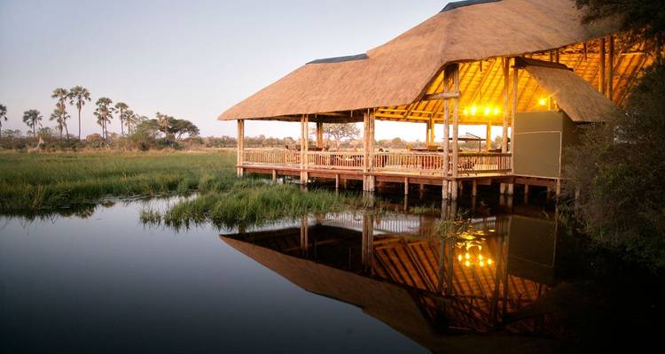 Un lodge de safari magnifiquement éclairé avec des lumières chaleureuses au bord de l'eau pendant le crépuscule.