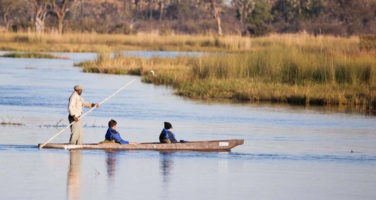 Un homme pagayant un canoë avec deux passagers sur une rivière sereine avec des herbes et des arbres.