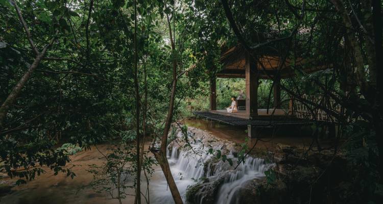 Vrouw zittend in meditatie op een houten paviljoen bij een waterval.