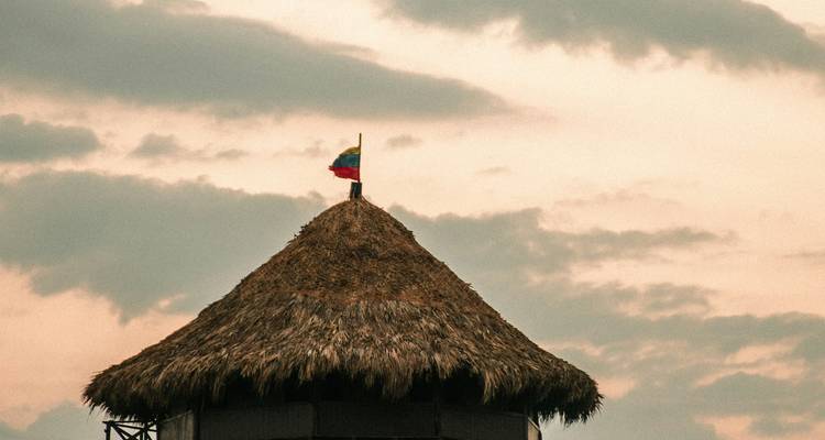 Techo de cabaña con techo de palma coronado por la bandera colombiana silueteada contra nubes de atardecer pastel