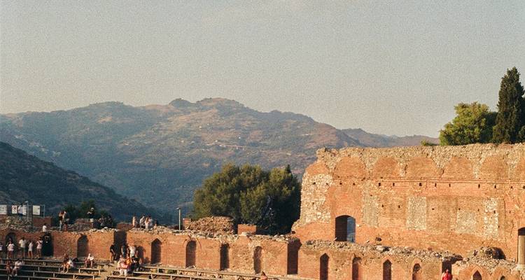 Las ruinas del antiguo teatro griego se alzan sobre las colinas ondulantes de Sicilia con visitantes explorando el sitio.