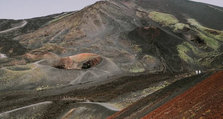 Los amplios cráteres volcánicos y las laderas de lava coloreada del Monte Etna con senderistas que empequeñecen en escala.