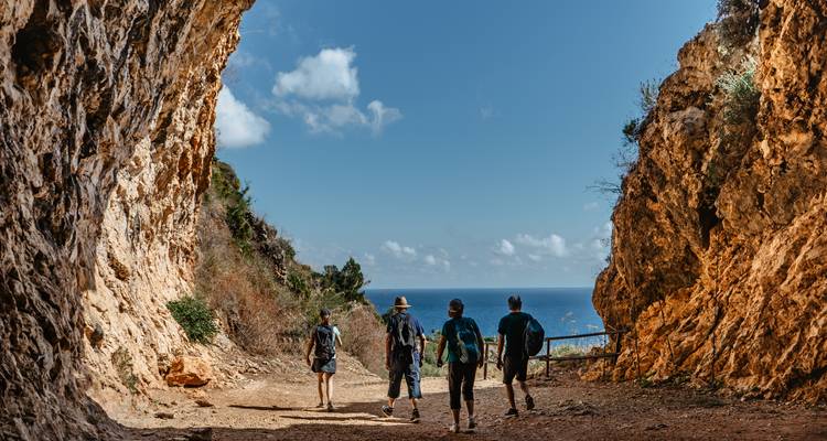Cuatro excursionistas salen de un sendero de cueva costera rocosa hacia el mar azul y el cielo en un día soleado.