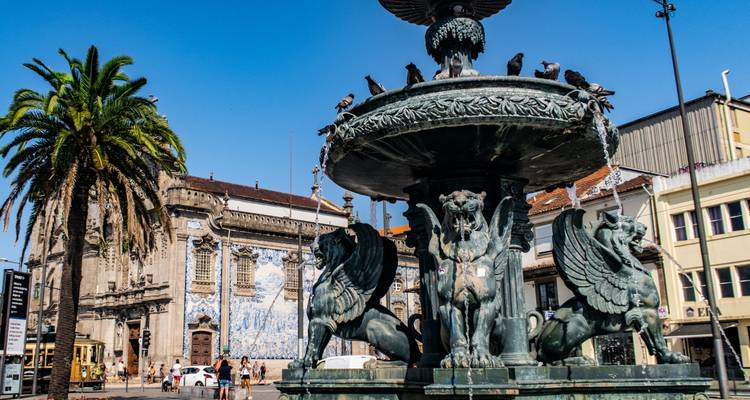 Fuente decorativa de bronce con criaturas mitológicas en una plaza cerca de una iglesia de azulejos azules en Oporto.