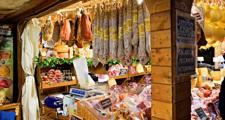 Puesto de mercado de delicatessen cubierto mostrando carnes curadas colgadas, quesos y productos locales.