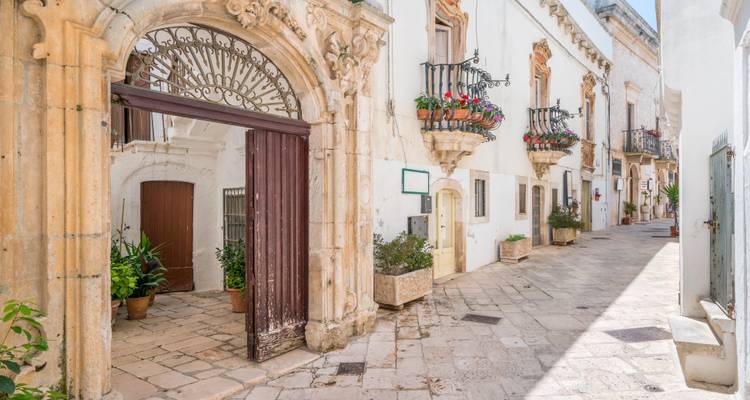 Elegante entrada barroca que abre a una calle de piedra iluminada por el sol, adornada con balcones y flores.