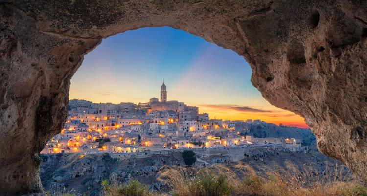 Vista del atardecer de las viviendas en cuevas iluminadas de Matera enmarcadas por un arco de roca natural.
