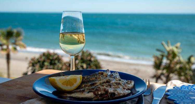 Un plato de sardinas a la parrilla y una copa de vino blanco se ponen frente a una vista del mar Mediterráneo de color turquesa.