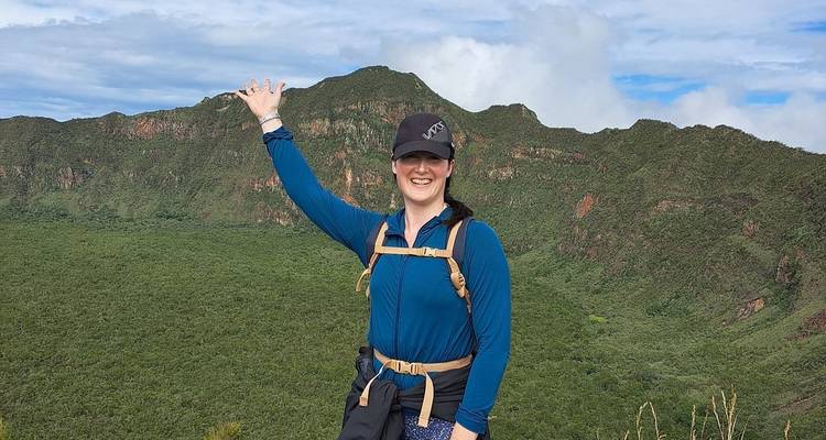 Person, die auf einem Wanderweg mit üppiger Berglandschaft im Hintergrund posiert.