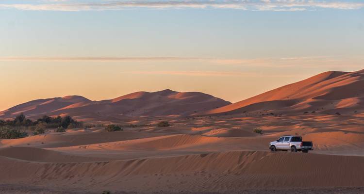 Weite Wüstenlandschaft mit einem Fahrzeug, das bei Sonnenuntergang durch Sanddünen navigiert.