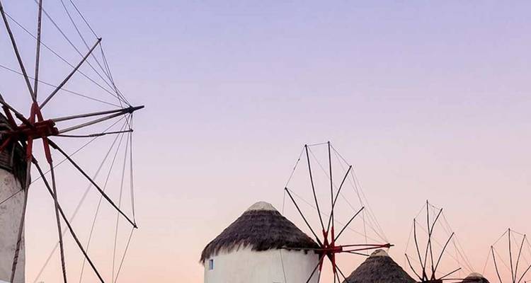 Silhouetten van klassieke witte windmolens met rode zeilen onder een zachte pastelkleurige avondhemel op Mykonos.