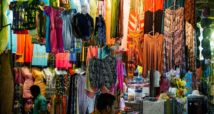 Scène de marché colorée avec des vêtements suspendus et divers textiles.