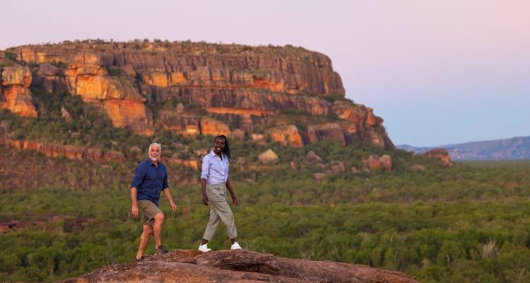 Een paar loopt bovenop een door de zon verlicht rotsplateau met een dramatische steilrand als achtergrond in Kakadu.