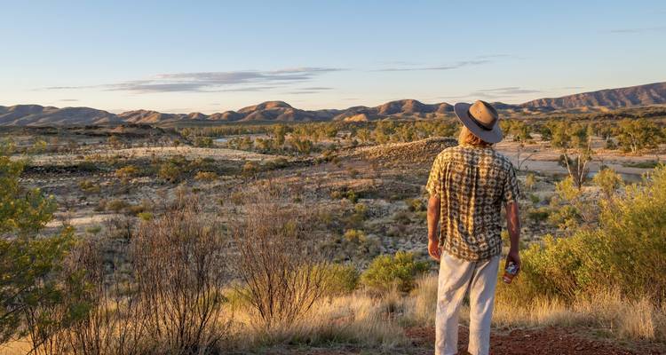 Voyageur au chapeau contemple la vaste vallée de l'Outback avec sa végétation clairsemée et ses chaînes de montagnes lointaines au coucher du soleil