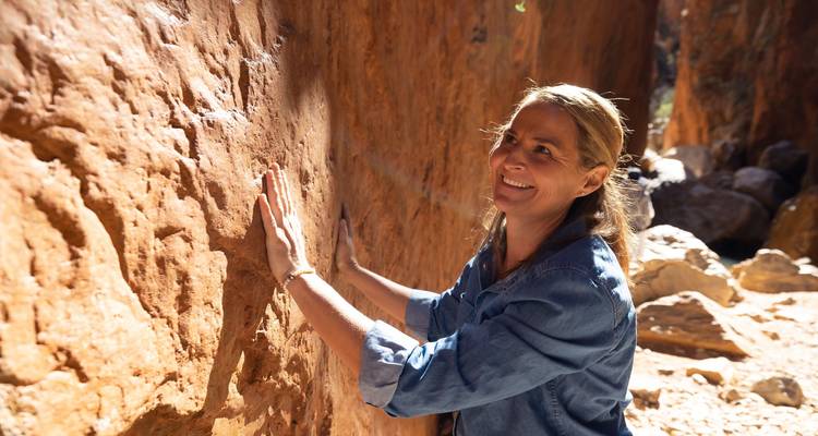 Femme souriante explorant une gorge étroite de roches rouges, la main sur un mur de grès texturé
