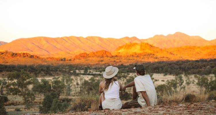 Couple assis sur un promontoire rocheux regardant la chaîne de montagnes briller d'orange dans la lumière du coucher de soleil