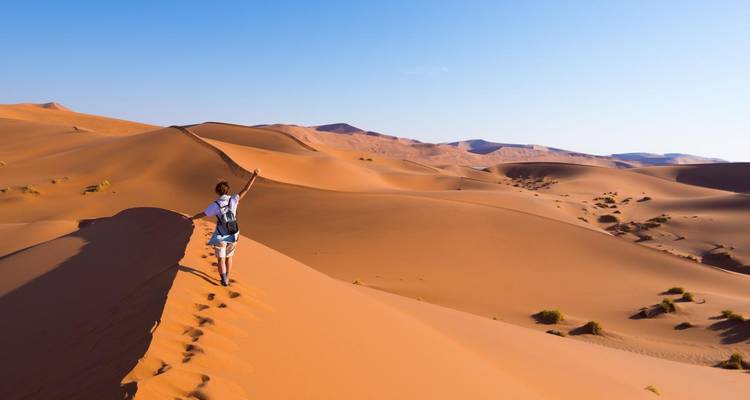 Un mochilero camina por la cresta de una enorme duna de arena naranja bajo un cielo azul despejado.
