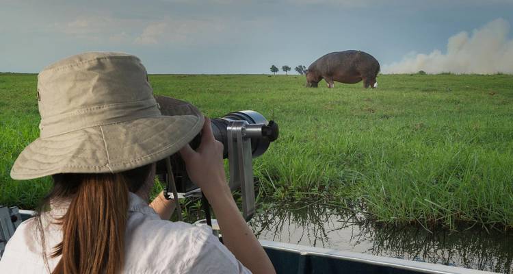 Viajero con lente de telefoto fotografía un hipopótamo que está comiendo pasto desde una pequeña barca en el agua.