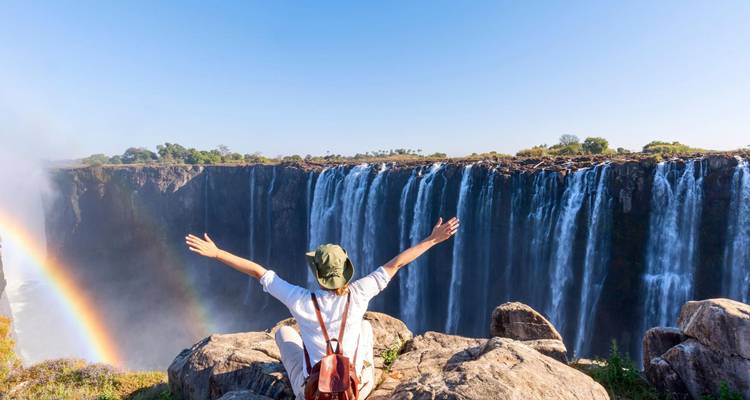 Viajero con los brazos extendidos celebra frente a las Cataratas Victoria y un arcoíris vibrante.