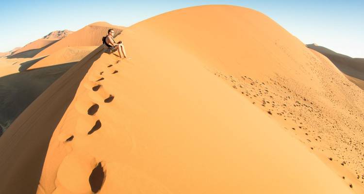 Un hombre descansa en la cima de una enorme cresta de duna de arena, con huellas que quedan atrás de él.