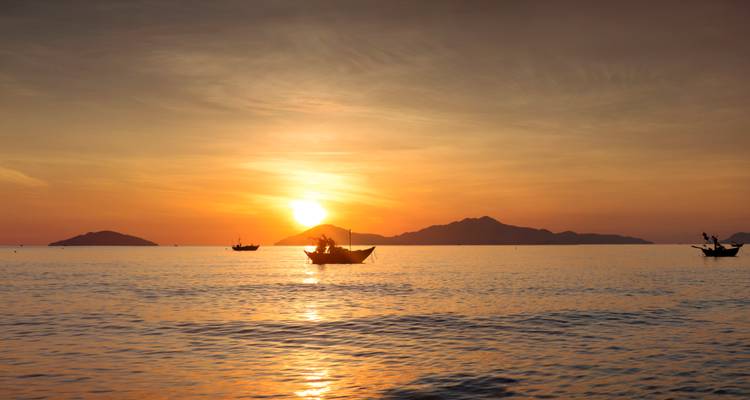 Amanecer dorado sobre el mar tranquilo con barcas de pesca y siluetas de islas.