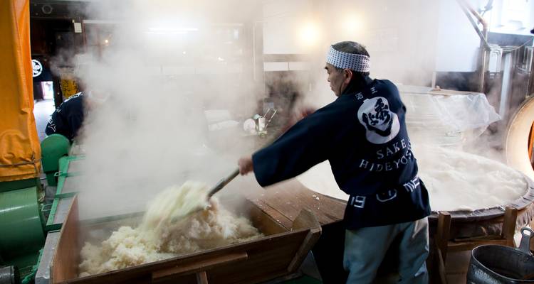Brewer stirs steaming rice mash during traditional sake production in a rustic workshop.