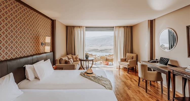 Elegant hotel room with twin beds opens onto a balcony overlooking a desert lake and hills.