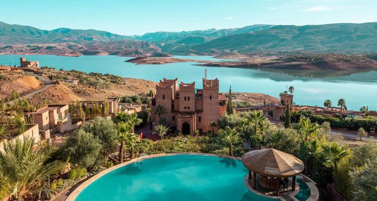 Hilltop kasbah resort with circular pool overlooks a vast turquoise lake and snow-dusted peaks.