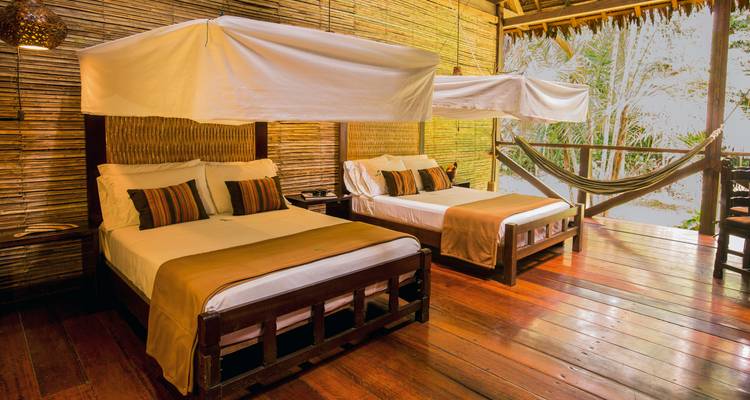 Comfortable eco-lodge room with two canopy beds, wooden floors and a hammock overlooking jungle foliage.