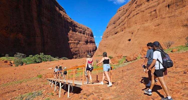 Los excursionistas cruzan un puente pequeño entre enormes paredes de arenisca roja bajo cielos azules brillantes.
