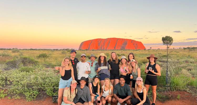 Groupe de touristes souriants posant au coucher de soleil avec le monolithe rouge lumineux d'Uluru en arrière-plan.