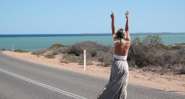 Una joven levanta los brazos triunfalmente mientras camina por una carretera costera con vistas a las aguas turquesas de Australia.