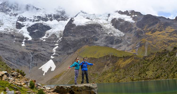 Two people posing by a lake with dramatic mountain scenery in the background.