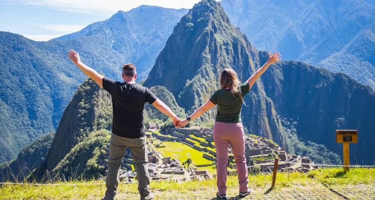 Een stel dat hand in hand loopt met hun armen hoog in de lucht kijkt uit over de iconische ruïnes van Machu Picchu, omlijst door dramatische Andespijlers.