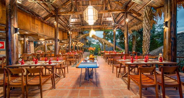 Restaurant tropical en plein air avec des poutres en bois, un toit en chaume et des tables soigneusement dressées, délicatement éclairé pour le dîner.