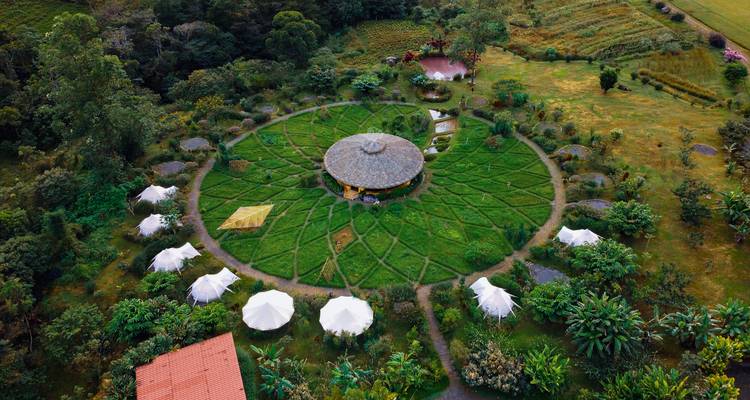 Drone view of circular eco-lodge with surrounding tented glamping pods set in lush gardens.