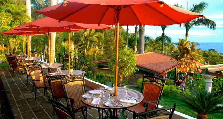 Outdoor restaurant terrace with red umbrellas overlooking tropical gardens and the sea.