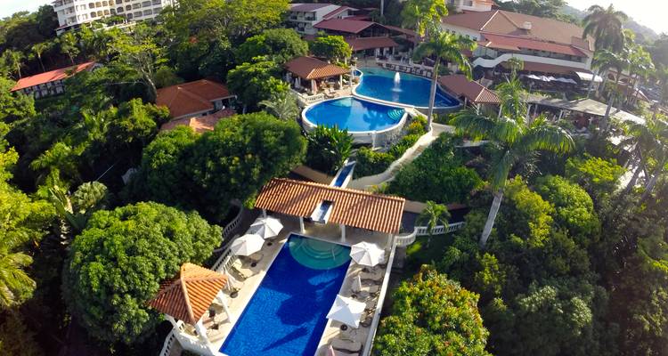 Aerial view of tiered blue swimming pools nestled among dense tropical trees at a hillside resort.