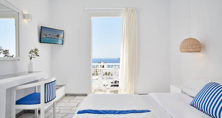 Habitación de hotel blanca y luminosa con una puerta de balcón que se abre a una vista del mar azul, muebles minimalistas y cojines azules acentuados.