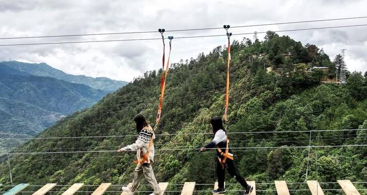 Two people walking on a suspension bridge in a forested mountainous area.