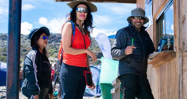 Hikers wearing hats and sunglasses check in at a wooden hut surrounded by Kilimanjaro base camp tents.
