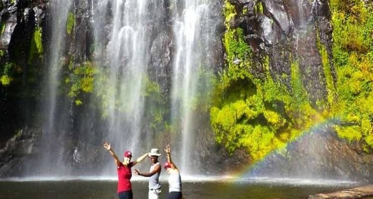 Three people celebrate beneath a tall jungle waterfall with a vivid rainbow in the spray.