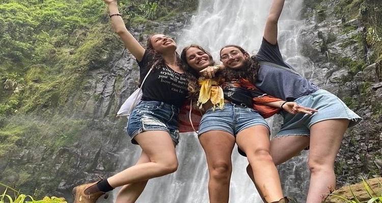 Three young women in shorts joyfully pose in front of a tall jungle waterfall surrounded by lush greenery.