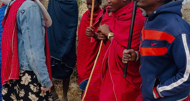 Local Maasai men in traditional shukas converse with travelers in a dry grassy landscape.