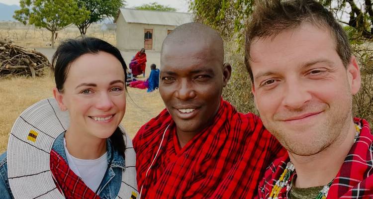 Smiling travelers pose with a Maasai local for a close-up selfie outside a rural dwelling.