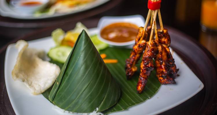 Traditional Indonesian food on a plate with rice cone and skewered meat.
