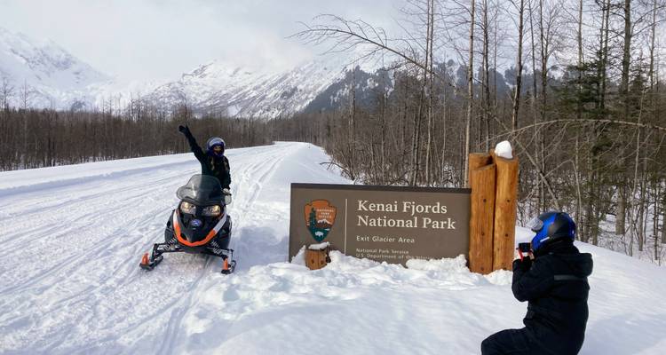 Sneeuwscooterrijder poseert bij het bord van Kenai Fjords National Park.